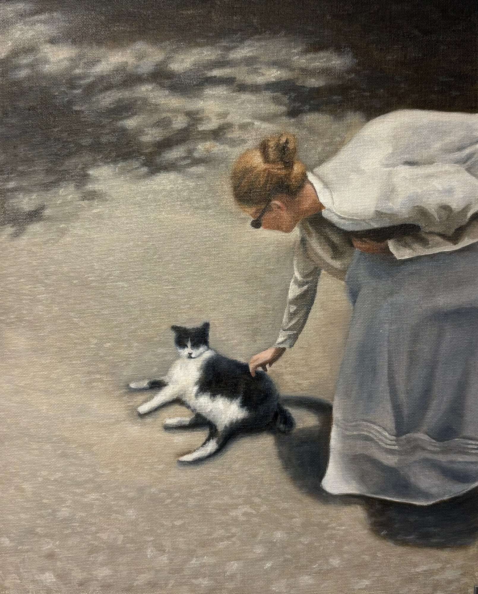 Seen from a high angle, a woman in a white blouse and long pale blue skirt bends down to stroke a black-and-white cat stretched out on sunlit ground, her shadow falling across the paving beside it.