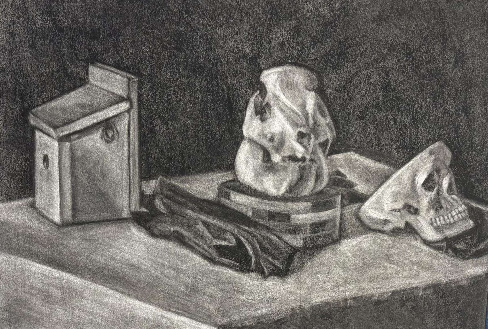 A charcoal still life of an animal skull and a human skull resting on a table beside a metal box and a draped cloth, lit against a dark ground.